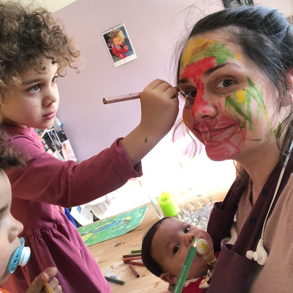 Eine Mutter mit einem Baby in der Trage wird von ihren beiden anderen Kindern mit Stiften bunt im Gesicht angemalt.